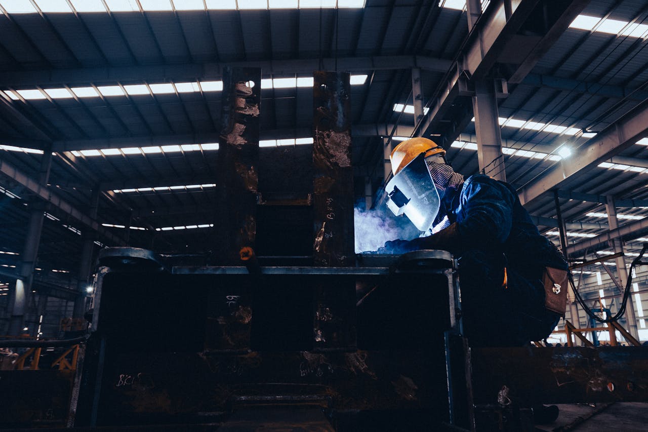 A welder wearing protective gear works on metal in a dimly lit industrial workshop.